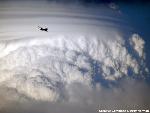 An airplane is circumnavigating a large cumulonimbus cloud. The airplane appears as bird compared to the size of the cloud. The top of the cloud is sheared off by winds.