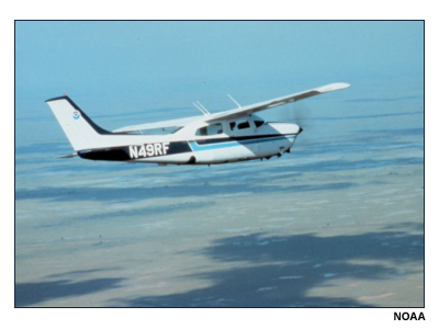 Photo of small airplane over flat terrain with cloud shadows below.