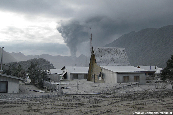 Casas sepultadas bajo ceniza volcánica, que llega a las ventanas del segundo piso, en un pueblo cerca del volcán Chaitén, en Chile. Se ve la columna eruptiva a lo lejos.